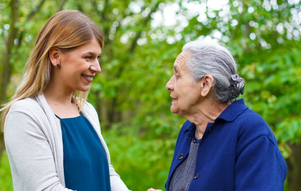 A happy caregiver supports and elderly woman walking outside trees in the background.