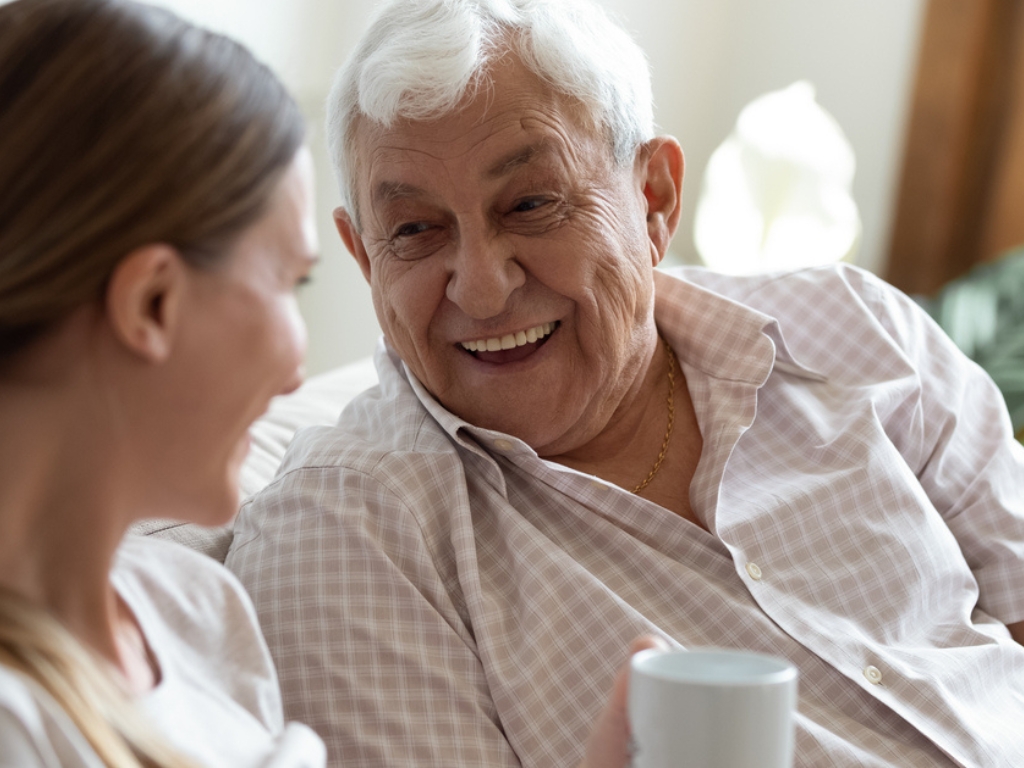 A elderly man laughs looking at a care giver next to him on a couch.