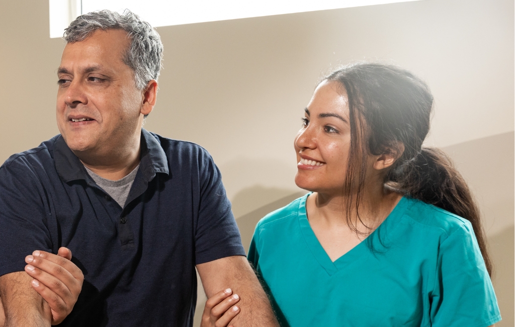 A caregiver in teal scrubs smiling warmly while assisting a senior man indoors, symbolizing compassionate senior home care in Palm Beach Gardens.