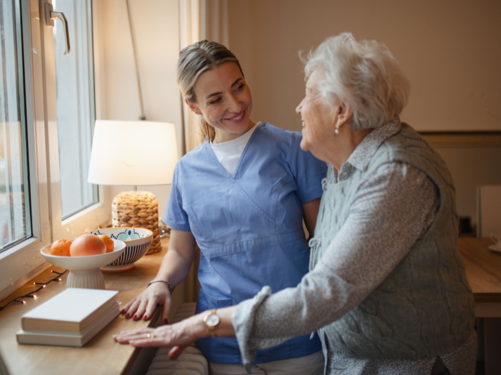 Caregiver providing home care in North Palm Beach, smiling warmly while supporting an elderly woman near a window, creating a sense of comfort and companionship.
