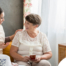 Smiling caregiver with her hand on an older woman’s shoulder as they share tea on the couch, showing home care vs assisted living