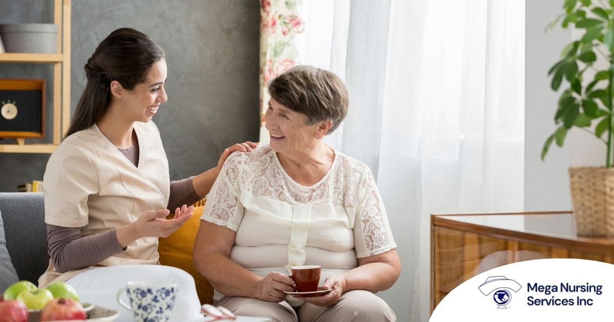 Smiling caregiver with her hand on an older woman’s shoulder as they share tea on the couch, showing home care vs assisted living